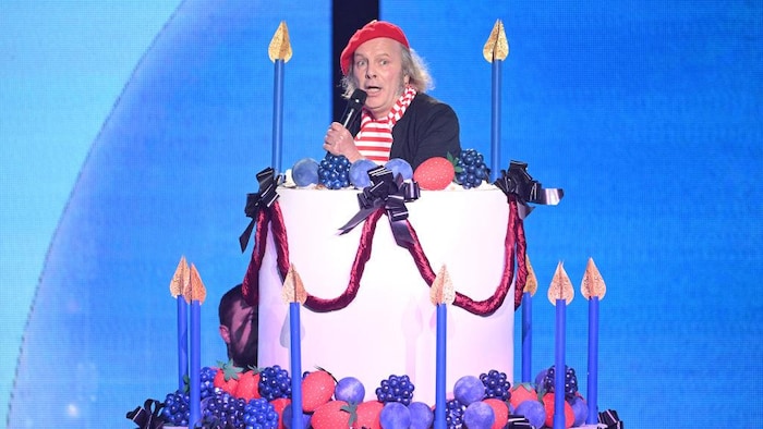 Un homme portant un béret rouge chante en sortant d'un gâteau de fête géant.
