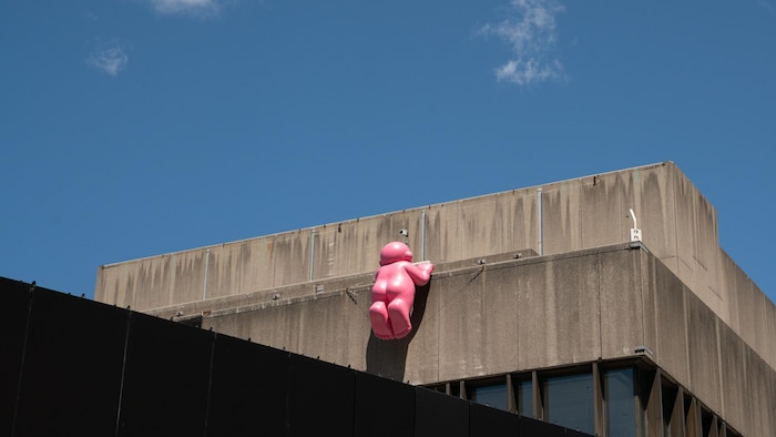 On voit un immeuble en béton découpé sur un ciel bleu. Une sculpture rose se tient sur la rambarde.
