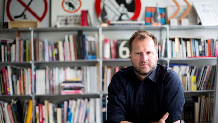 Un homme prend la pose, devant une bibliothèque garnie de livres et d'objets. 