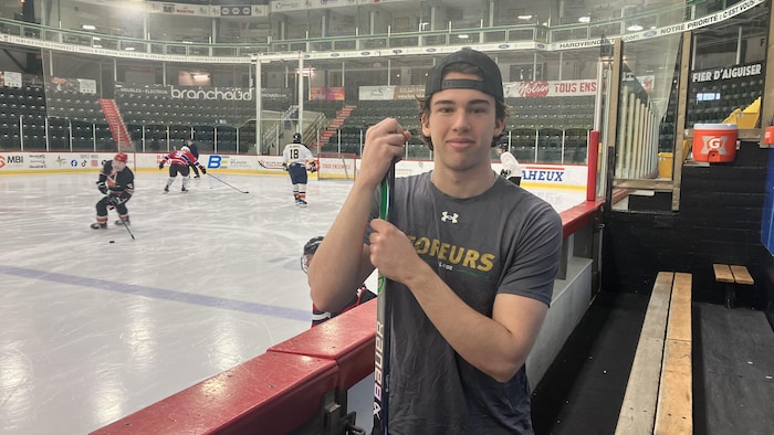 Philippe Veilleux, casquette sur la tête et appuyé sur un bâton de hockey au banc de son équipe, pose pour la caméra en souriant légèrement.