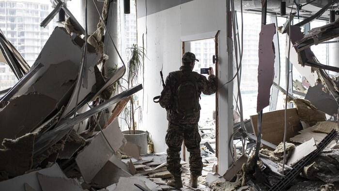 Un homme en habit militaire prend une photo avec son téléphone dans sa main droite. Dans sa main gauche, il tient une arme.