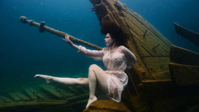 Une femme sous l'eau, près d'une épave, sa jambe droite allongée vers l'avant.