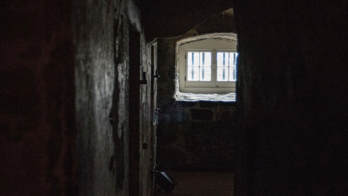 Vue de l'entrée de la prison au sous-sol du Morrin Centre. Un long couloir sombre et étroit mène à une pièce où se trouvent une petite fenêtre ainsi que des portes de cachots.