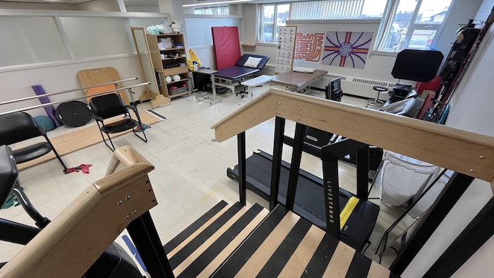 Une salle de réadaptation physique avec un petit escalier de bois, un tapis roulant et des tables de traitement.