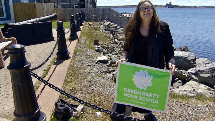 Clare Brett avec dans les mains une pancarte du Parti vert. Elle est près de la rive. 