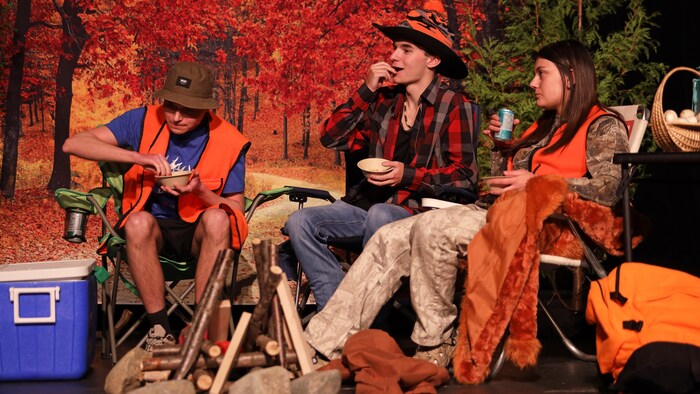 Trois jeunes acteurs assis sur des chaises pliantes devant un décors forestier sur une scène.