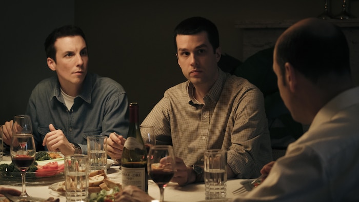 Deux jeunes hommes regardent leur père à table. 