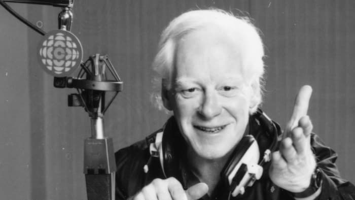L'animateur Pierre Bourgault, des écouteurs encerclant son cou, est assis à une table où reposent ses notes qu'il tient d'une main. Près de sa tête, on voit un micro suspendu qui porte une affichette avec le logo de Radio-Canada.