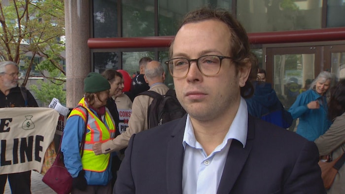 Pierre Chauvin porte une chemise et un veston. Derrière lui, une dizaine de personnes, certaines tiennent une pancarte. Ils se trouvent tous devant un bâtiment.