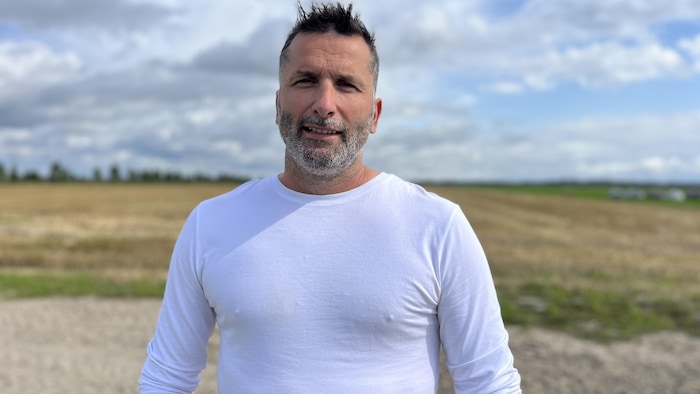 Un homme avec un chandail blanc pose devant un champ.