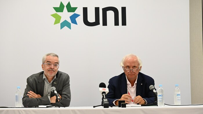 Pierre-Marcel Desjardins and Camille Theriault sat next to each other, leaning at a table, during a press conference.  Above their heads is the UNI crest. 