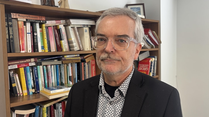 Une personne portant des lunettes regarde la caméra. Il se trouve devant une bibliothèque remplie de livres sur l'économie.