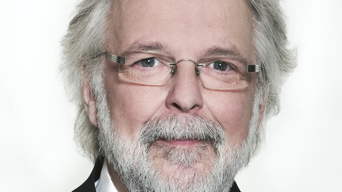 Un homme aux cheveux blancs, à la barbe blanche, il porte des lunettes