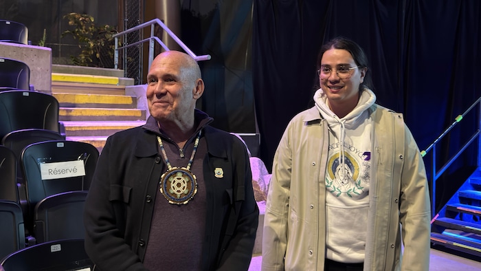 Un homme souriant et portant une médaillon perlé accompagné d'un plus jeune homme qui sourit également.
