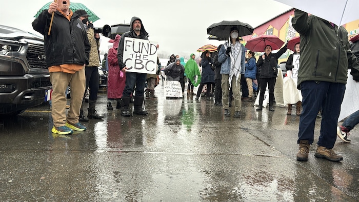 Des manifestants sous la pluie. Sur une pancarte, on lit «Fund the CBC».