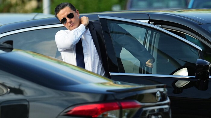 Un homme avec des lunettes de soleil en train de porter une veste à sa sortie d'une voiture.