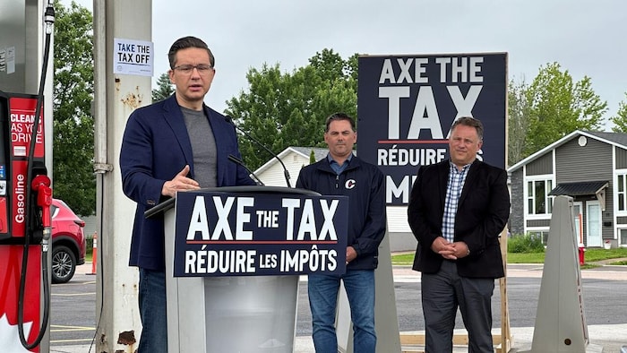 Pierre Poilievre parle devant un lutrin et une affiche où est écrit « Axe the Tax », posée à côté des pompes à essence d'une station-service.