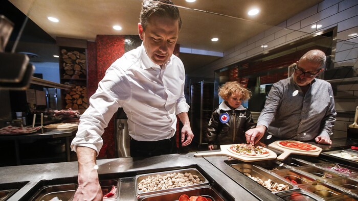 Pierre Poilievre prépare une pizza avec lad fils et un homme à côté.