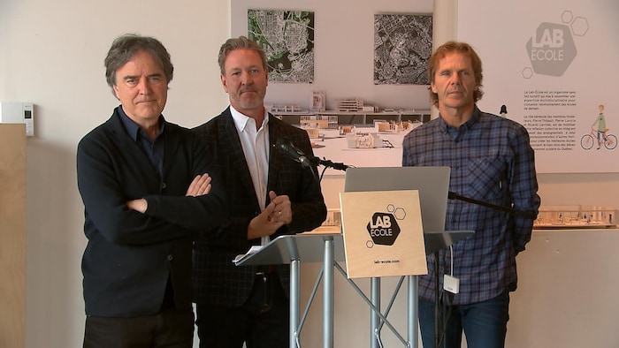 Trois hommes debout devant un lutrin sur lequel est posé le rapport du Lab-École avec, en arrière-plan, des maquettes.