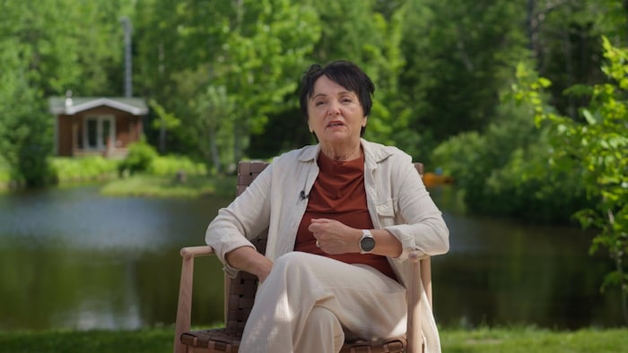 Une femme aux cheveux courts foncés est assise sur une chaise devant un petit lac.