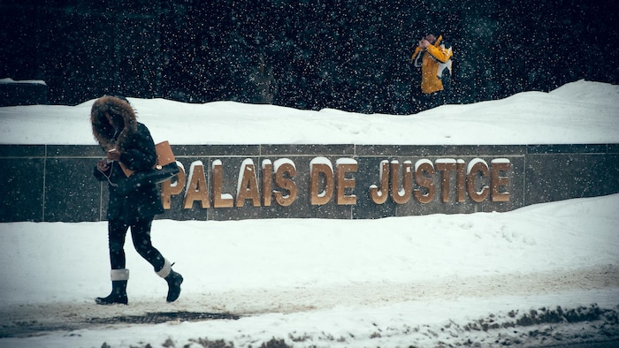 Une femme passe devant l'enseigne du palais de justice de Montréal, sous une fine neige. Un homme s'allume une cigarette à l'entrée de l'édifice.