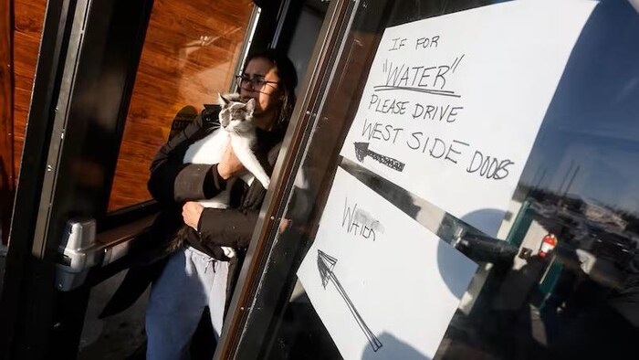 Une femme qui porte un chat dans ses bras sort d'un immeuble. Une affiche faite à la main indique aux résidents de Pimicikamak qu'ils doivent se rendre à la porte ouest pour obtenir de l'eau, le 7 janvier 2026.