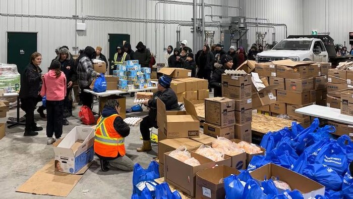 Des résidents dont la file dans une grande salle où se trouvent des boites et des sacs contenant des aliments, à Pimicikamak, le 7 janvier 2026.
