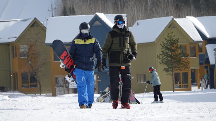 Deux planchistes marchent, équipés contre le froid, au pied de la station de ski Pin Rouge.