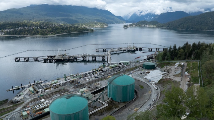 Une vue aérienne du point d'arrivée du pipeline Trans Moutain. Deux grandes cuves vertes et des structures de tuyaux au bord d'une baie entourée de montagnes. 