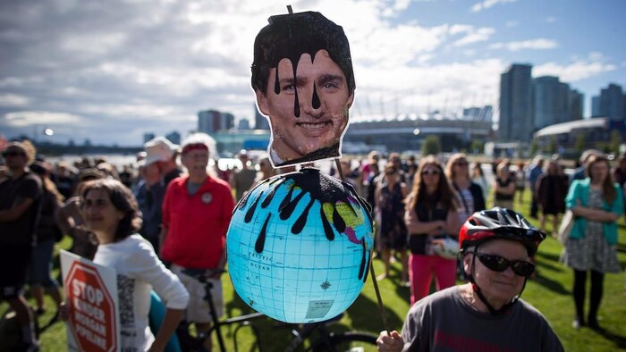 Un homme âgé tient un globe terrestre avec le visage de Justin Trudeau couvert en partie de pétrole noir. Des manifestants sont à l'arrière-plan devant un centre-ville.