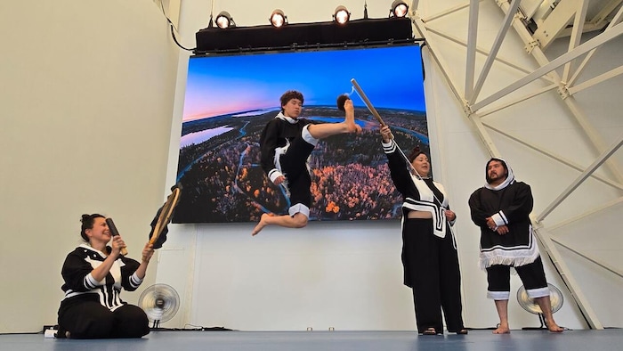 Un jeune homme saute pour toucher un bâton lors d’une démonstration de jeu inuk à l’Expo d’Osaka, À gauche, une femme est assise et tient un tambour, tandis qu’une autre femme et un homme en tenue traditionnelle assistent à la scène.