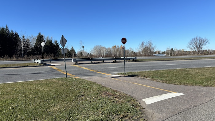 Une piste cyclable asphaltée mène à la traverse d'une large route bordée de glissières de sécurité.