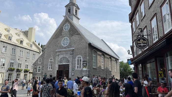 Une foule de touristes se massent devant une vieille église de pierres, et à la terrasse d'un restaurant situé à proximité.