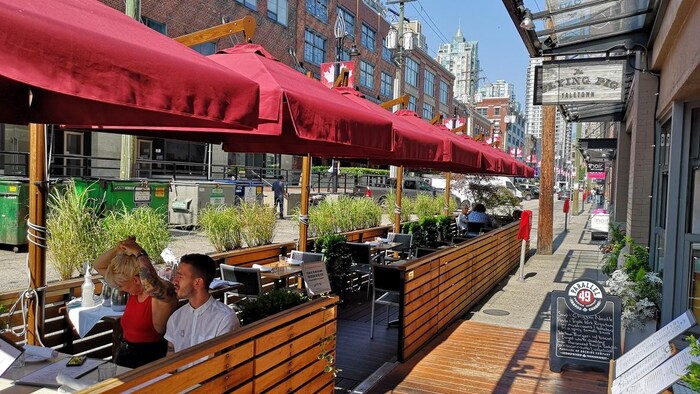 À droite, un restaurant, à gauche, une terrasse en bois avec des parasols qui empiète dans la rue, où on voit deux clients. En arrière, on voit des plantes qui la séparent de la rue.