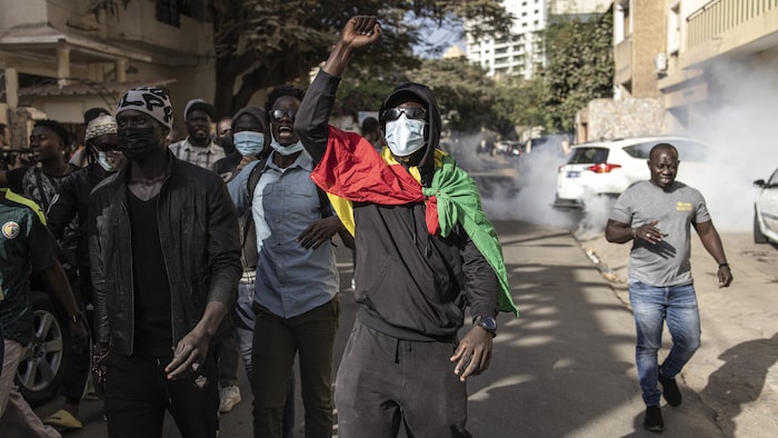 Des manifestants masqués déambulent dans une rue du quartier Plateau au Sénégal. De la fumée émerge d'une voiture garée en bordure.