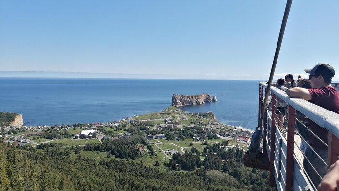 Des touristes sont appuyés sur les bords d'une plateforme qui donne une vue sur le village de Percé et le rocher Percé.