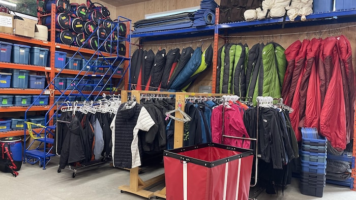 Des dizaines de sacs de couchage, matelas, vêtements d'extérieur et divers bacs sont rangés dans un entrepôt.