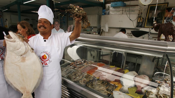 Un homme moustachu coiffé d'une toque de chef cuisinier lève sa main gauche qui tient un homard vivant et tient un gros poisson plat mort dans sa main droite.