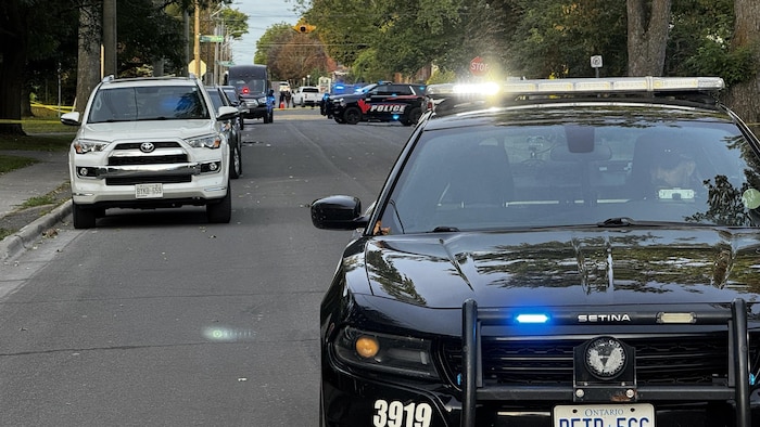 Une voiture de police en avant plan bloque une rue. D'autres véhicules sont garés sur la rue.