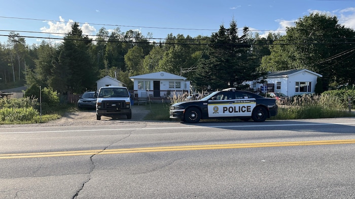 Un véhicule de la Sûreté du Québec est garé au bord d'une route devant des maisons blanches entourées d'arbres à Ragueneau.