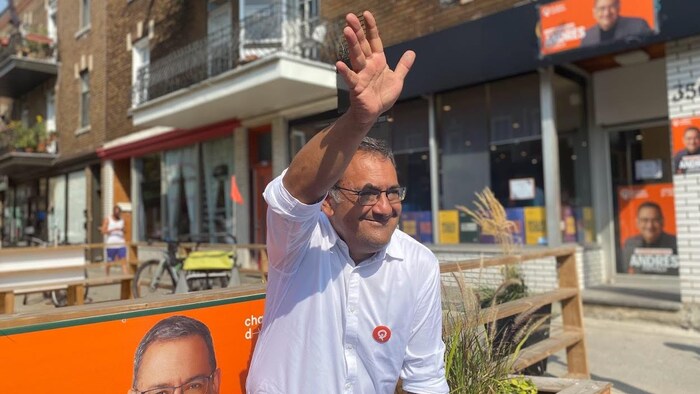 Un homme assis sourit, la main levée.