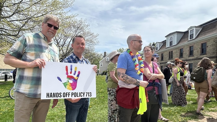 Des dizaines de personnes debout dehors devant l'Assemblée législative du Nouveau-Brunswick. Un homme exhibe un écriteau sur lequel est écrit « Hands Off Policy 713 ».