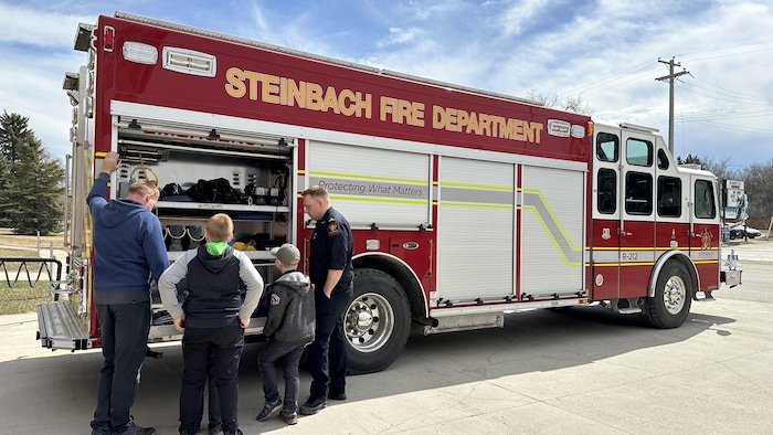 Des enfants, un père et un pompier sont debout devant un tiroir ouvert d’un camion de pompier. 