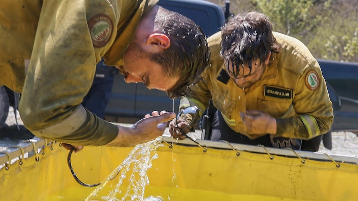 Deux pompiers à l'uniforme couvert de suie sont debout devant une grande cuve et s'aspergent le visage d'eau.