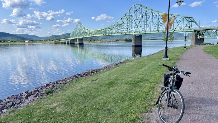 Un sentier pédestre le long d'une rivière, un vélo stationné, le pont en arrière plan.