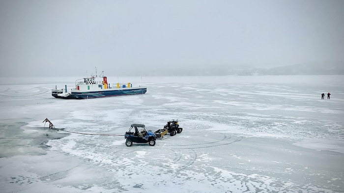 Un bateau pris dans la glace, des quatre-roues sur le lac gelé et deux silhouettes au centre du lac avec des piquets. 