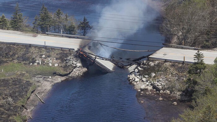 Un pont fumant, sous lequel on voit des flammes, est à moitié effondré dans un cours d'eau.