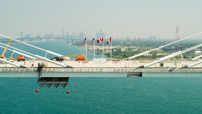 Le pont en construction, de la machinerie lourde sur le tablier. 2 drapeaux canadiens et 2 drapeaux américains flottent au vent chacun de leur côté de la frontière.