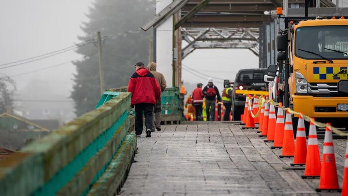 Le passage piétonnier d'un pont, où se trouvent d'un côté des passants et des employés de construction. De l'autre côté, des cônes et des camions.
