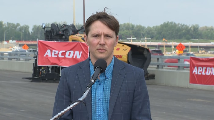 Devin Dreeshen s'adresse à la presse devant un microphone sur un chantier de construction routière, avec de la machinerie lourde en arrière-plan.